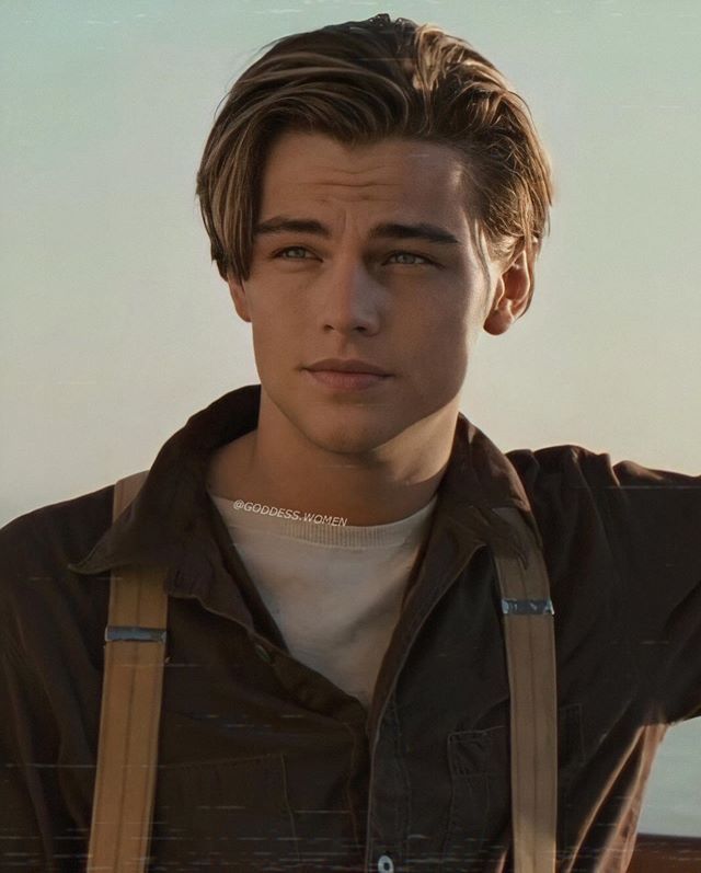 a young man with suspenders and a jacket looks at the camera while holding his hands on his head