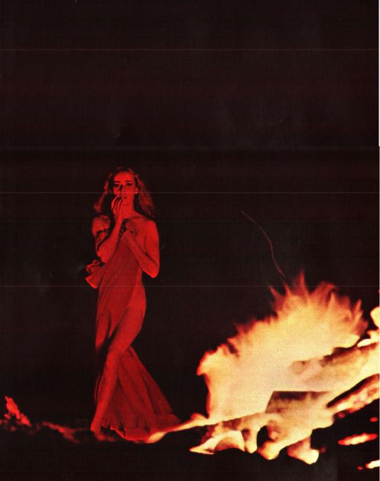 a woman in a red dress standing next to a fire with her hand on her face