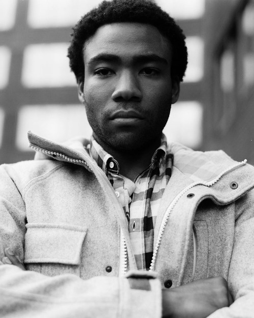 a man with his arms crossed standing in front of a brick building wearing a jacket