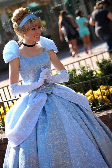 a woman in a blue dress and white gloves is smiling at someone on the street