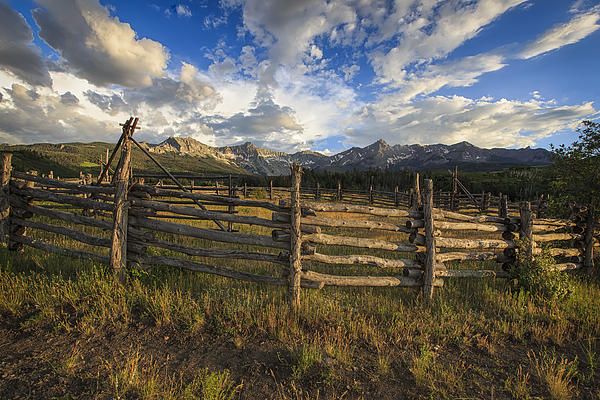 a wooden fence in the middle of a grassy field with mountains in the back ground