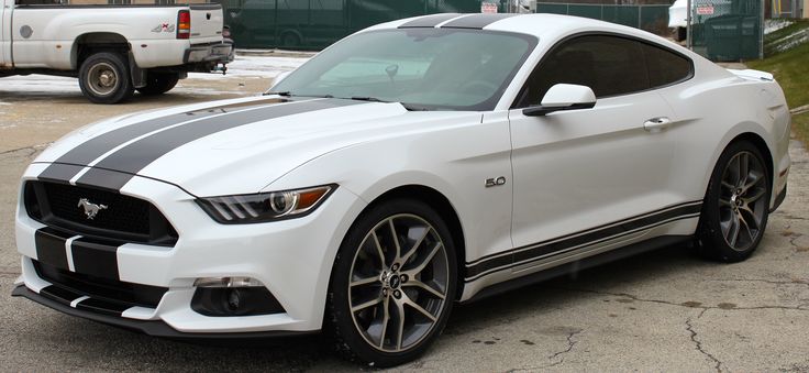 a white mustang with black stripes parked in front of a truck on the side of the road