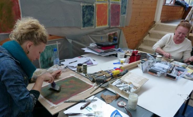 three women are sitting at a table with art supplies and paintings on the table in front of them