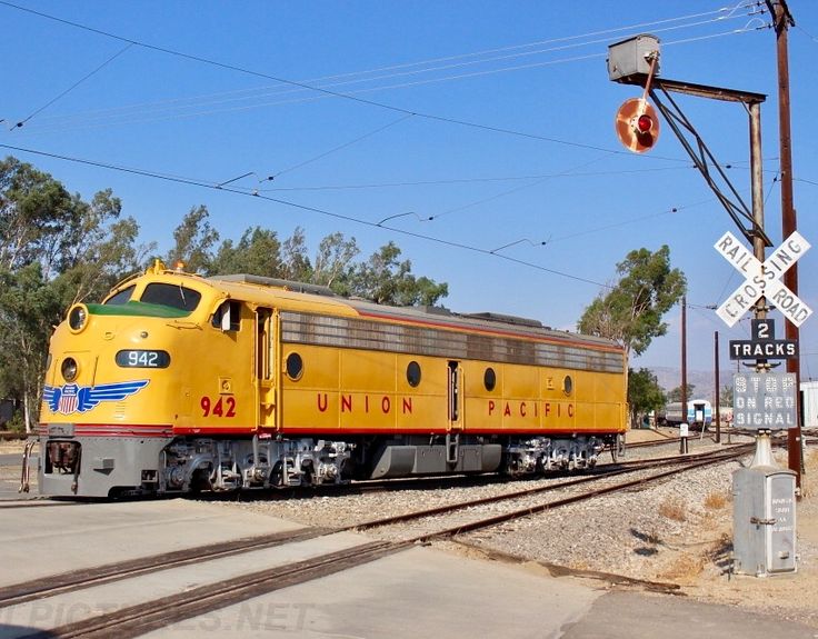 a yellow train traveling down tracks next to a railroad crossing sign and power lines with trees in the background