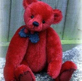 a red teddy bear with a blue bow sitting on top of a stone slab in front of a wooden fence