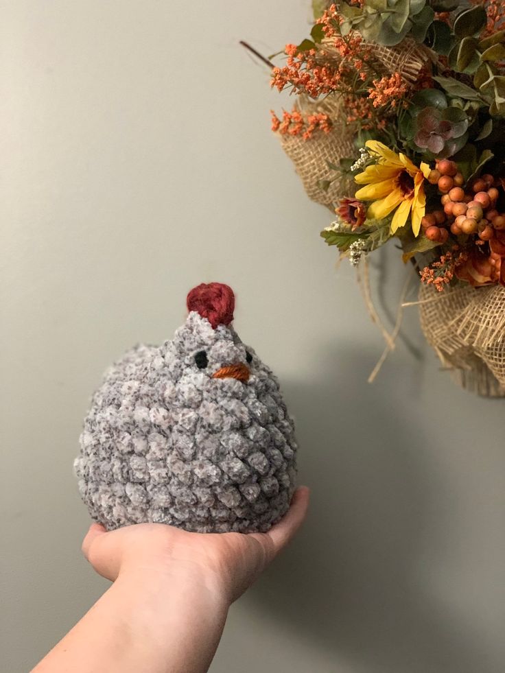 a hand holding a small crocheted chicken ornament next to a wreath