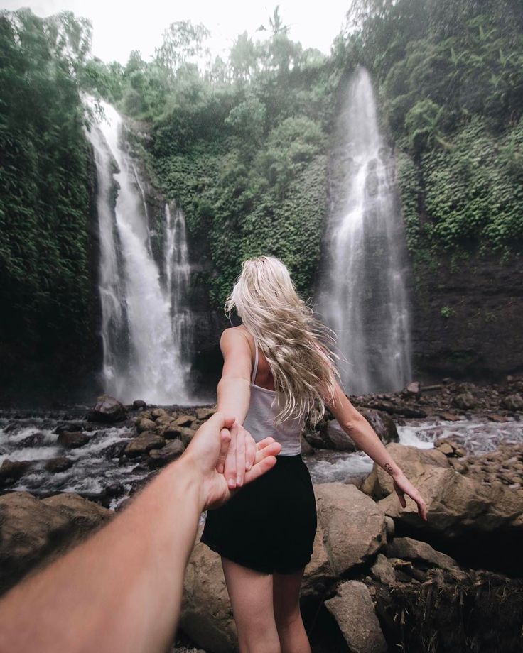 a person holding the hand of another persons hand in front of a waterfall with water cascading over it