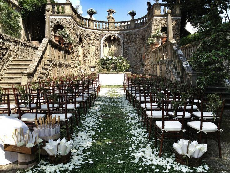 an outdoor ceremony set up with white flowers and petals on the aisle, in front of a stone building