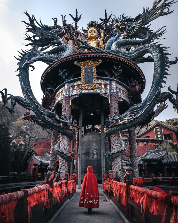 a woman in a red cape is walking towards a dragon statue at the entrance to a temple