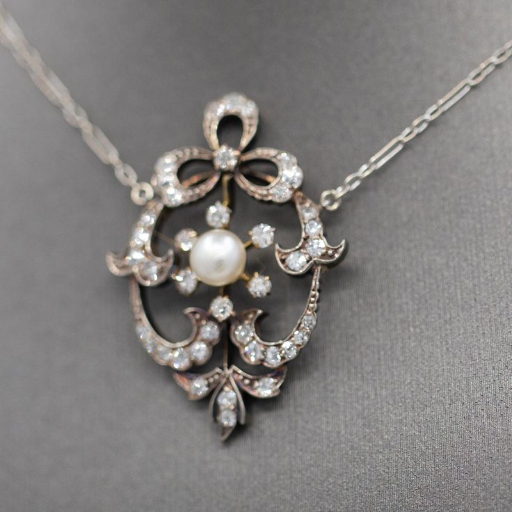 This is an absolutely exquisite necklace, beautifully crafted and replete with period details. The necklace chain is crafted in 14k white gold while the pendant is crafted in silver topped 14k yellow gold. The center natural pearl measures 5.5mm and is creamy white with lovely luster. The rest of the design is encrusted with roundish chunky old cut diamonds. The approximate carat weight is 0.74cttw. The diamonds are bright white and lively, no eye visible inclusions. The chain measures 16" and t Victorian White Gold Jewelry With Pearl Pendant, Vintage White Necklace With Pearl Pendant, Luxury Vintage Pendant Pearl Necklace, Ornate White Pearl Pendant Jewelry, Luxury Victorian Pearl Necklace With Pendant, Diamond Bows, Bow Necklace, Silver Tops, Natural Pearl