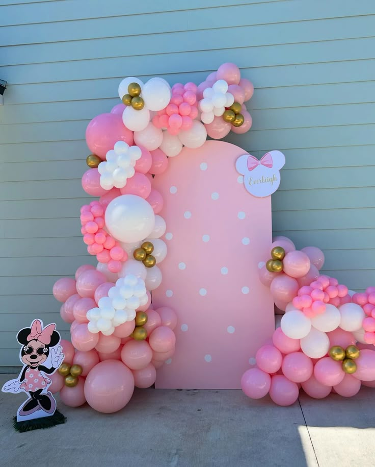 a pink and white balloon arch with minnie mouse balloons on it's side, next to a mickey mouse cake