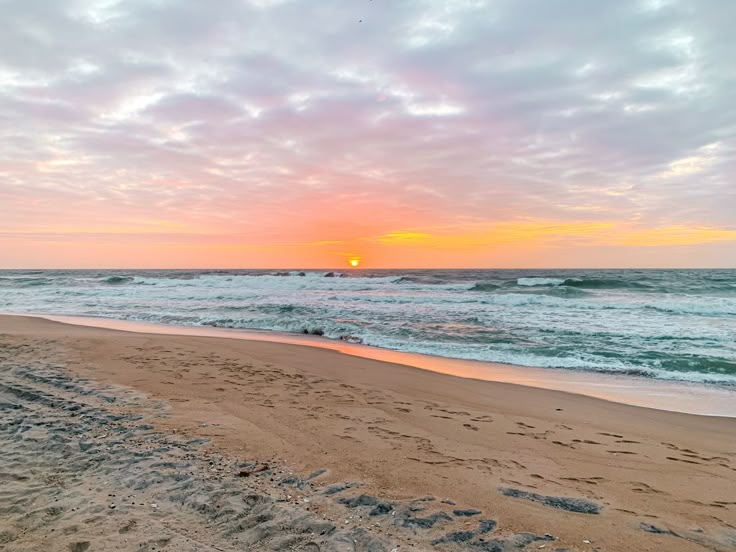 the sun is setting over the ocean with waves crashing on the beach and footprints in the sand