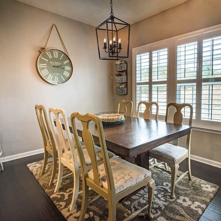 a dinning room table with chairs and a clock on the wall above it in front of two windows