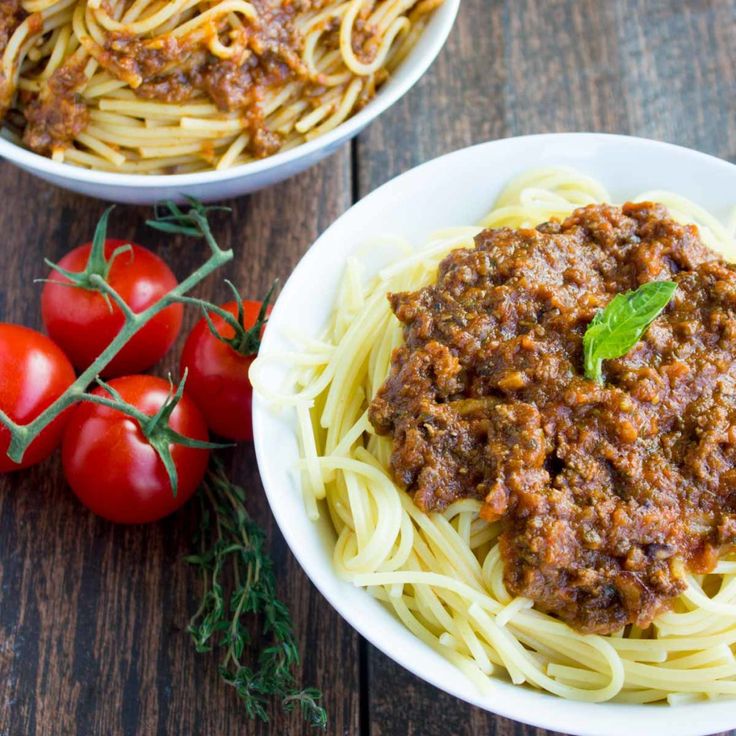 two white bowls filled with spaghetti and sauce next to tomatoes on a wooden table top