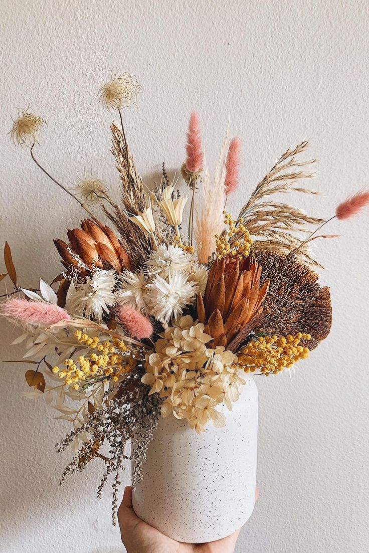 a hand holding a white vase with dried flowers and plants in it on a table