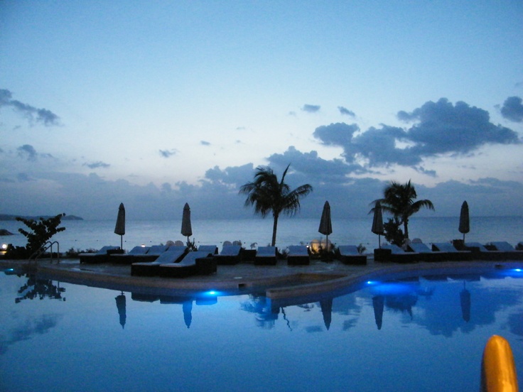 an empty swimming pool with lounge chairs and umbrellas next to the ocean at dusk