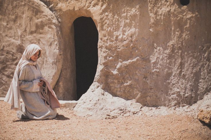 a woman sitting in front of a rock formation with a small cave behind her on the ground