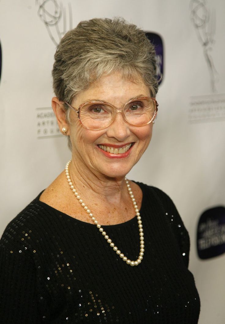 an older woman with glasses and pearls on her neck smiles at the camera while standing in front of a white backdrop