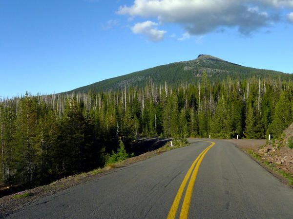 an empty road in front of a mountain with trees on both sides and blue sky above