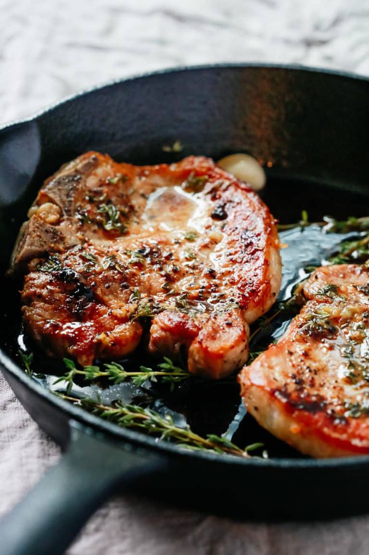 two pieces of meat in a skillet with herbs on the side and one piece being cooked