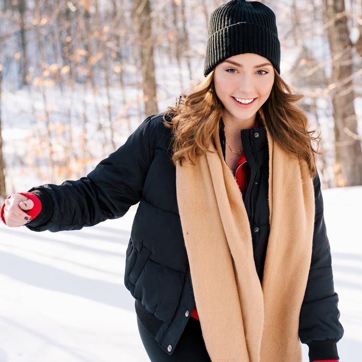 a woman in black jacket and brown scarf standing on snow covered ground next to trees