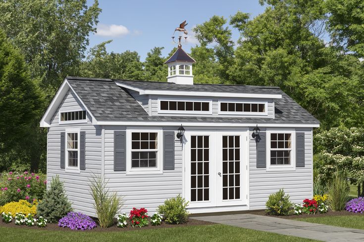 a small gray shed sitting in the middle of a yard with flowers and trees around it