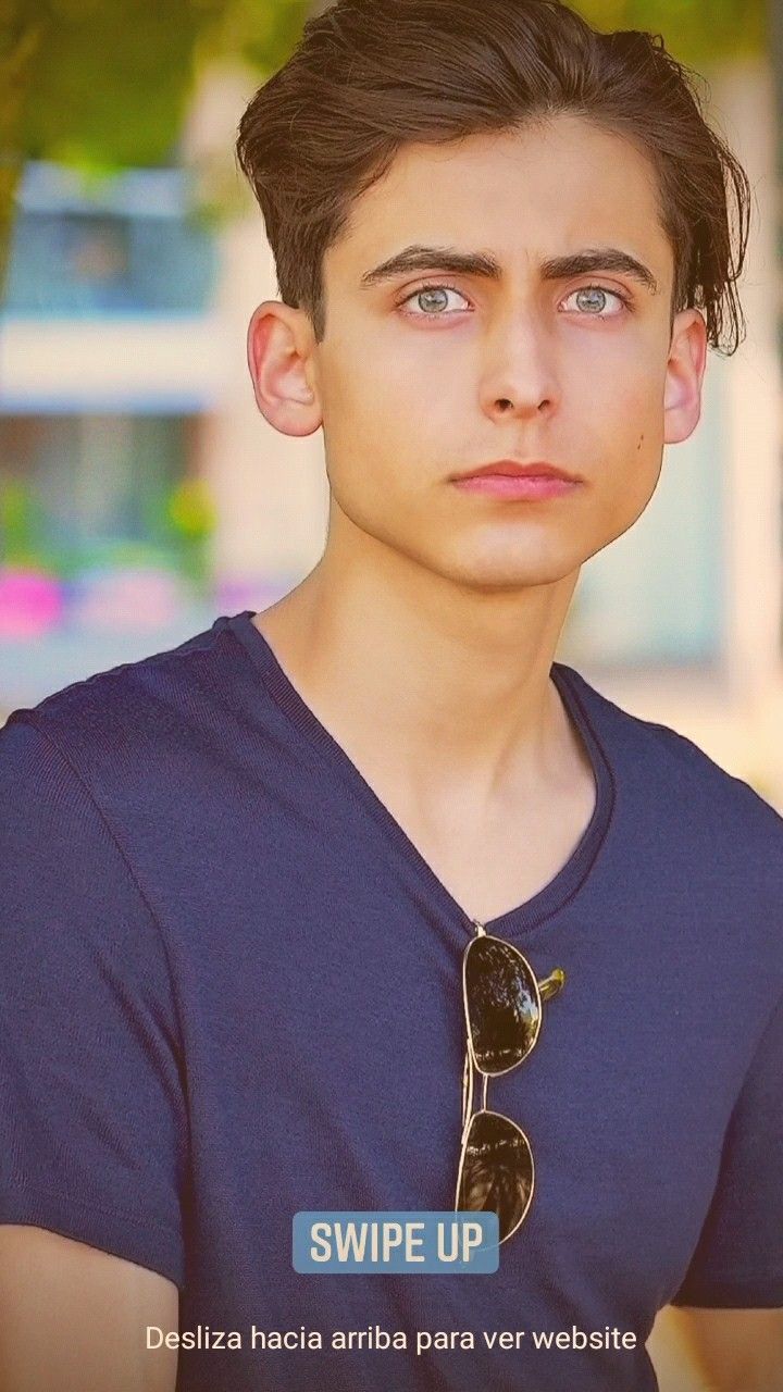 a young man with sunglasses on his neck looks at the camera while standing in front of a tree