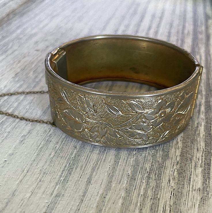 Add a touch of vintage elegance to your outfit with this beautiful Vintage Brass Hinged Bangle. The bracelet features a classic nouveau-style design with delicate floral etching, all in a stunning gold color. Approximately 1 inch tall About 2.5 inches wide Comes with original safety chain This hinged bangle is meticulously crafted and perfect for wearing alone as a statement piece or pairing with your favorite accessories. Don't miss out on adding this charming vintage piece to your collection! Elegant Antique Gold Bracelet Jewelry, Elegant Antique Gold Metal Bracelets, Antique Gold Adjustable Bracelets For Formal Occasions, Elegant Antique Gold Metal Bracelet, Antique Adjustable Cuff Bracelet For Formal Occasions, Elegant Wedding Bracelets With Vintage Charm, Vintage Hinged Bracelets For Wedding, Vintage Hinged Cuff Bracelet, Vintage Cuff Bracelet With Intricate Design For Formal Events