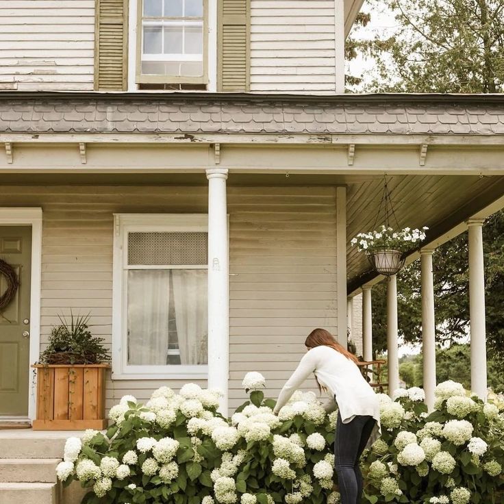 a woman standing on the front steps of a house next to hydrant plants and flowers