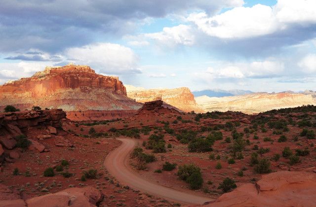 a dirt road going through the desert with mountains in the background and clouds above it