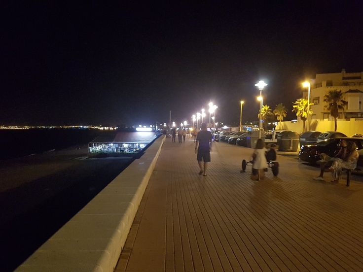 people are walking on the boardwalk at night near some parked cars and palm trees in the distance