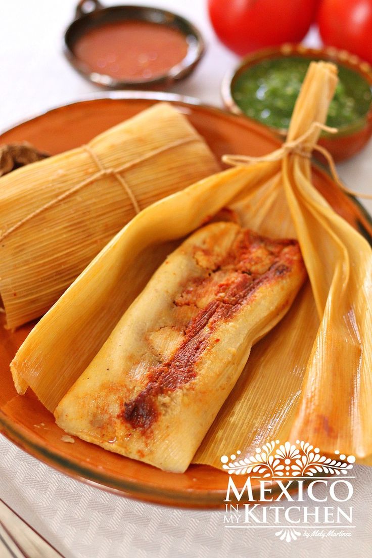 two tamales on a plate with dipping sauces in the background, and another item wrapped around it