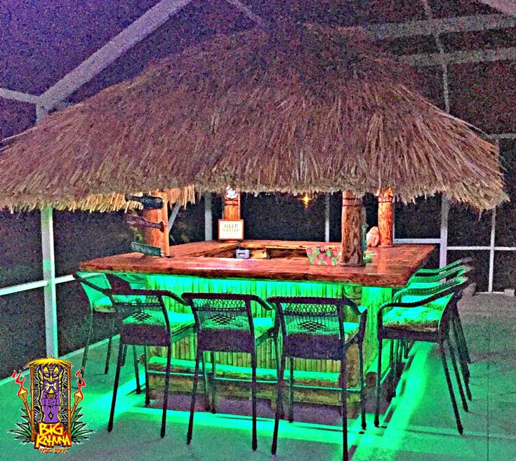 an outdoor bar is lit up with green lights and straw umbrellas on the roof