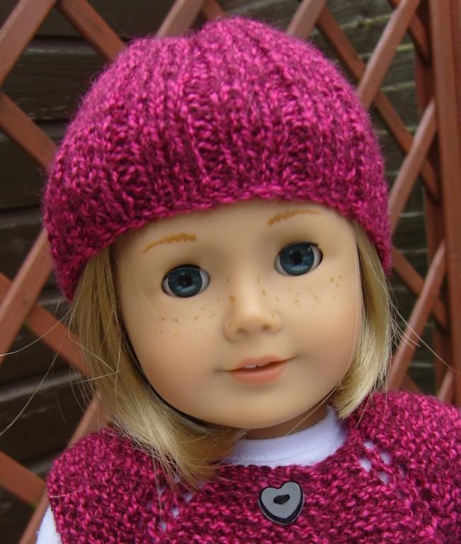 a doll with blonde hair wearing a pink knitted sweater and hat, standing in front of a wooden fence