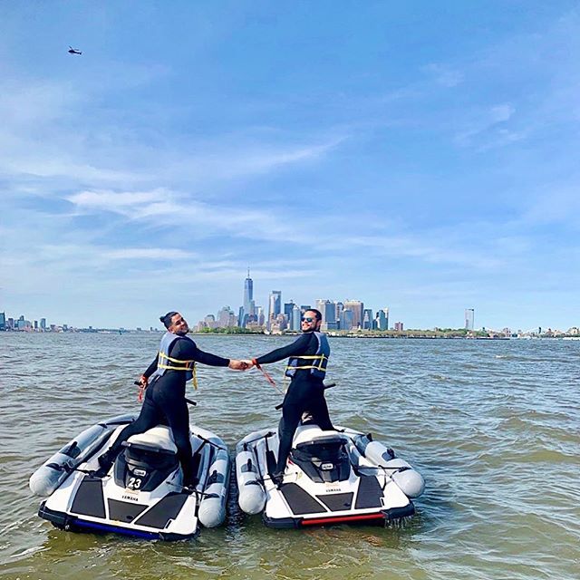 two men in wetsuits on jet skis shaking hands over the water with a city skyline in the background