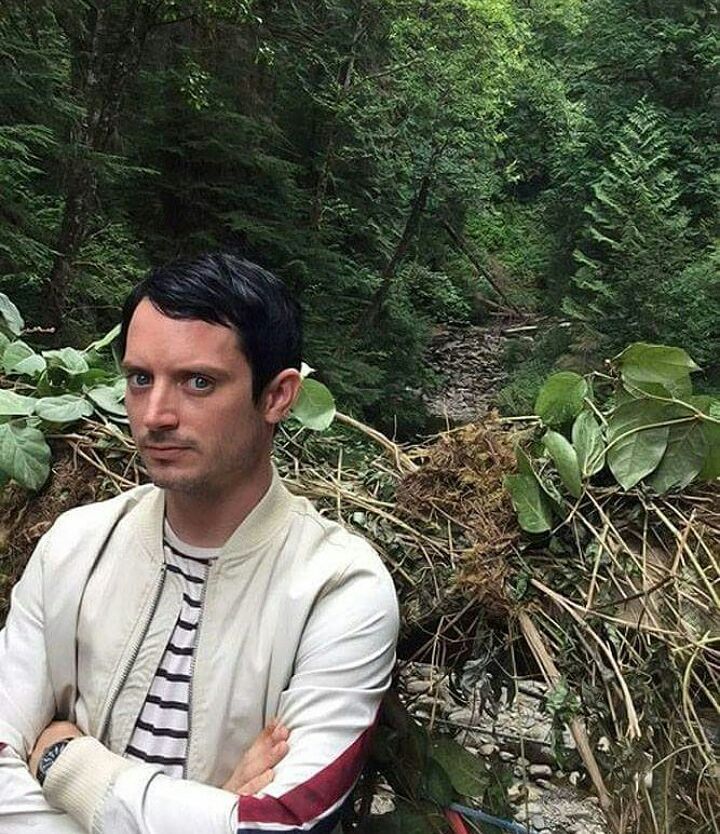 a man with his arms crossed standing in front of a stream surrounded by trees and bushes