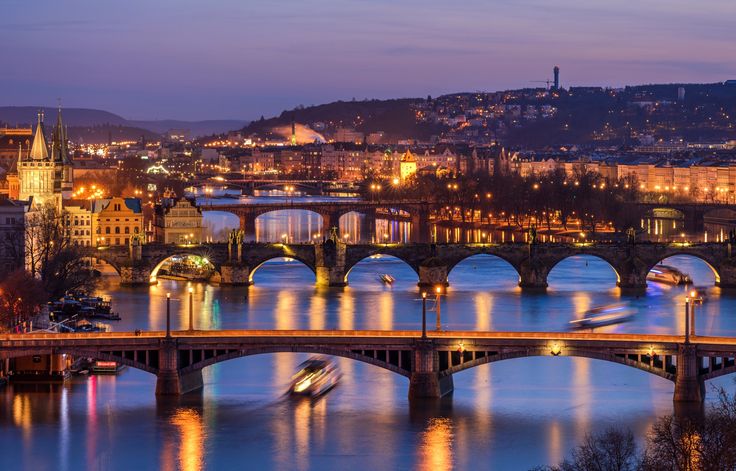 a bridge that is over some water with buildings in the background and lights on it