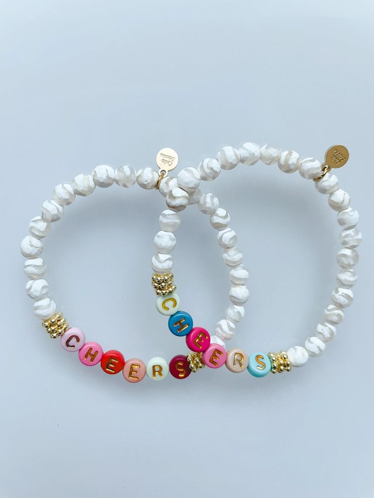 two white bracelets with colorful beads and gold charms on each beaded wristband