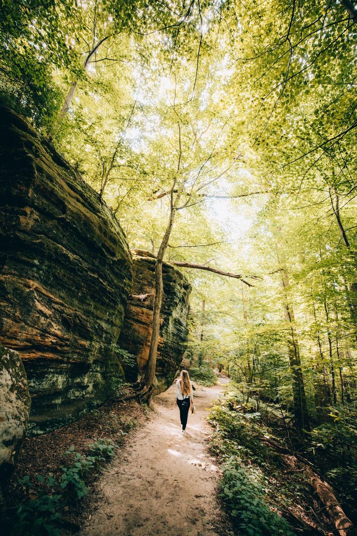 a woman walking down a dirt path through a forest filled with tall rocks and trees