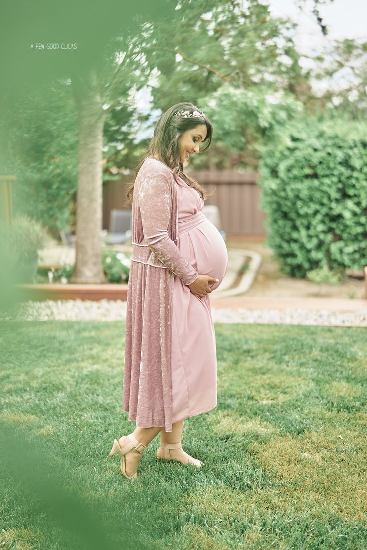 a pregnant woman in a pink dress is standing on the grass with her hands behind her back