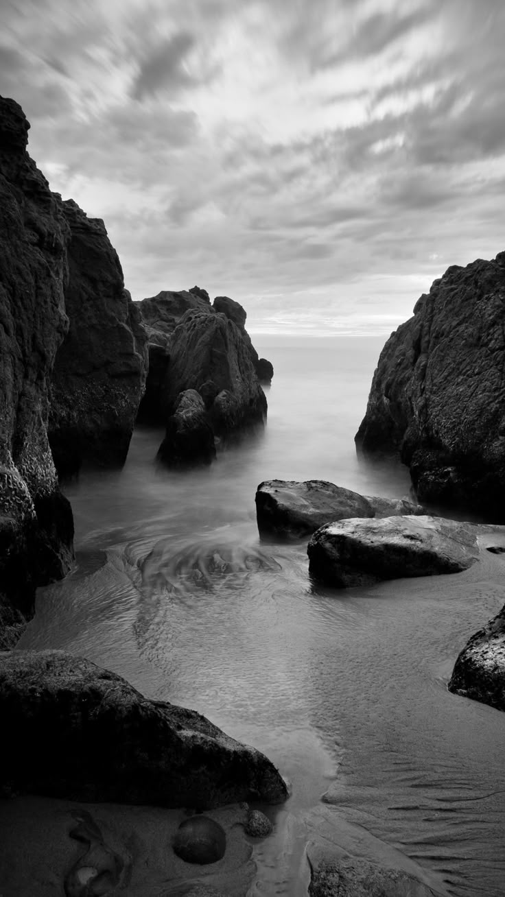 black and white photograph of rocks in the ocean with cloudy sky above them, along shore line