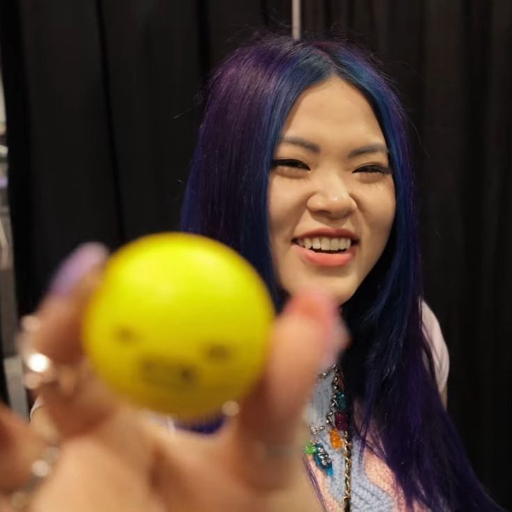 a woman with purple hair is holding an orange in her hand and smiling at the camera