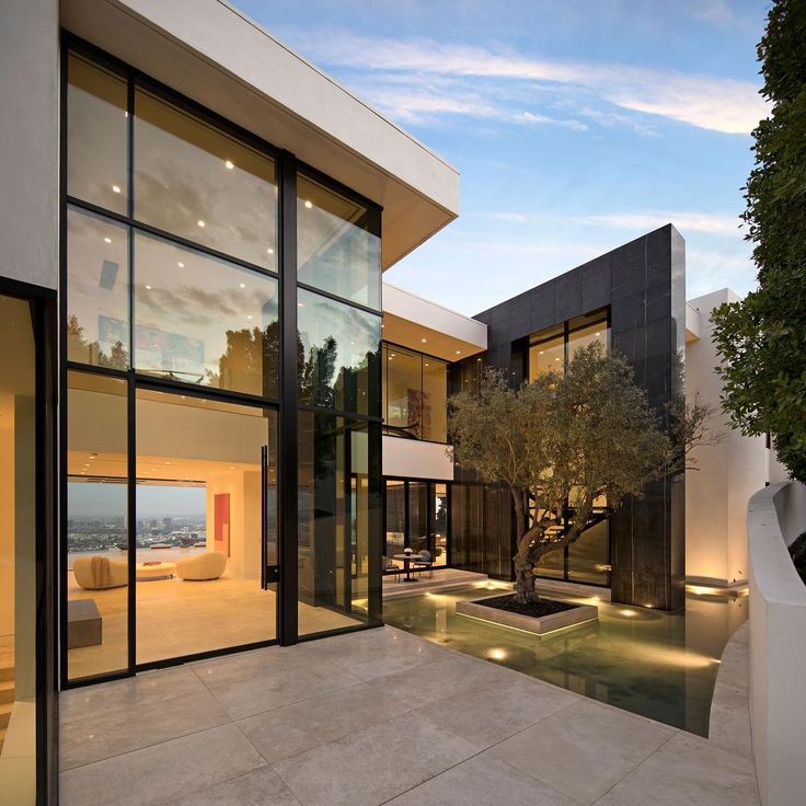 a modern house with large glass windows and an olive tree in the front yard at dusk