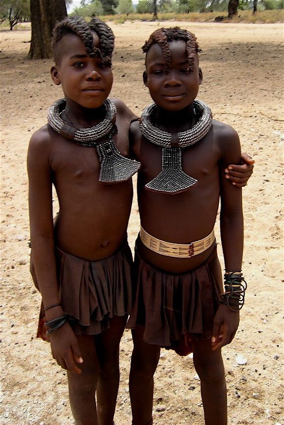 two young african children standing next to each other on a dirt ground with trees in the background