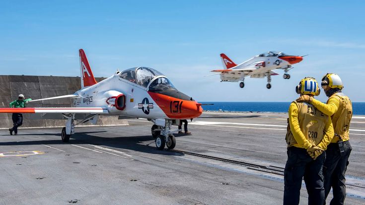 two men standing on an aircraft carrier looking at a fighter jet taking off from the flight deck