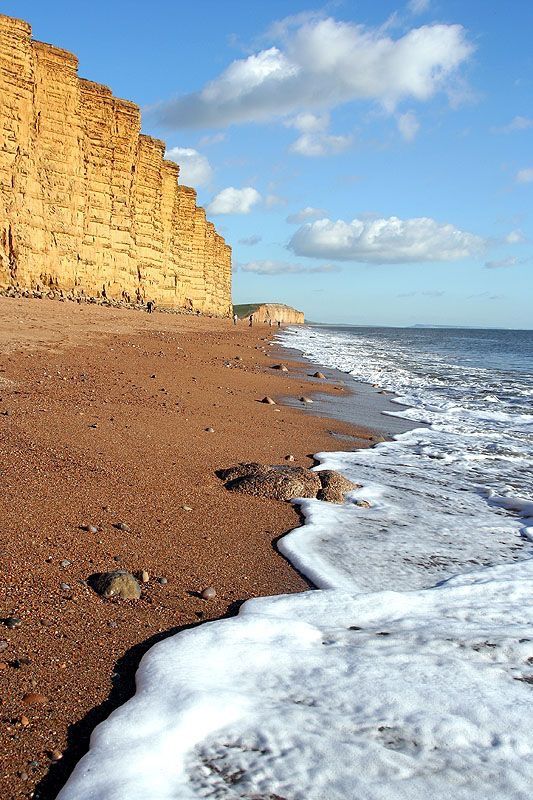 footprints in the sand on a beach next to water and cliff side with blue sky