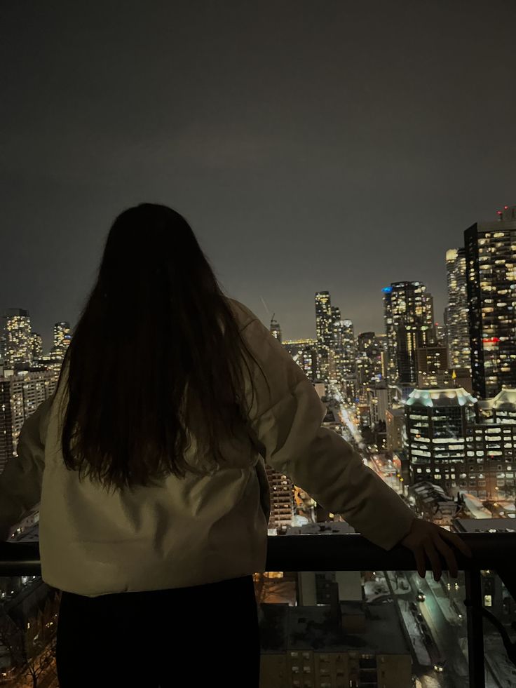 a woman standing on top of a tall building looking at the city lights in the distance