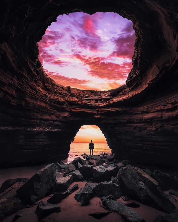 a person standing in the middle of a cave with rocks and water below it at sunset