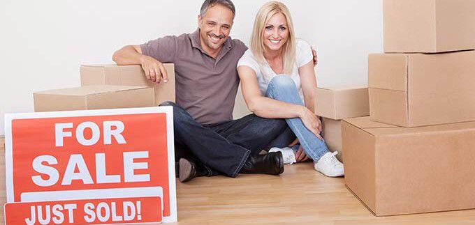 two people sitting on the floor with boxes and a for sale sign