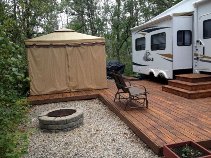 a camper trailer is parked next to a deck with a chair and fire pit